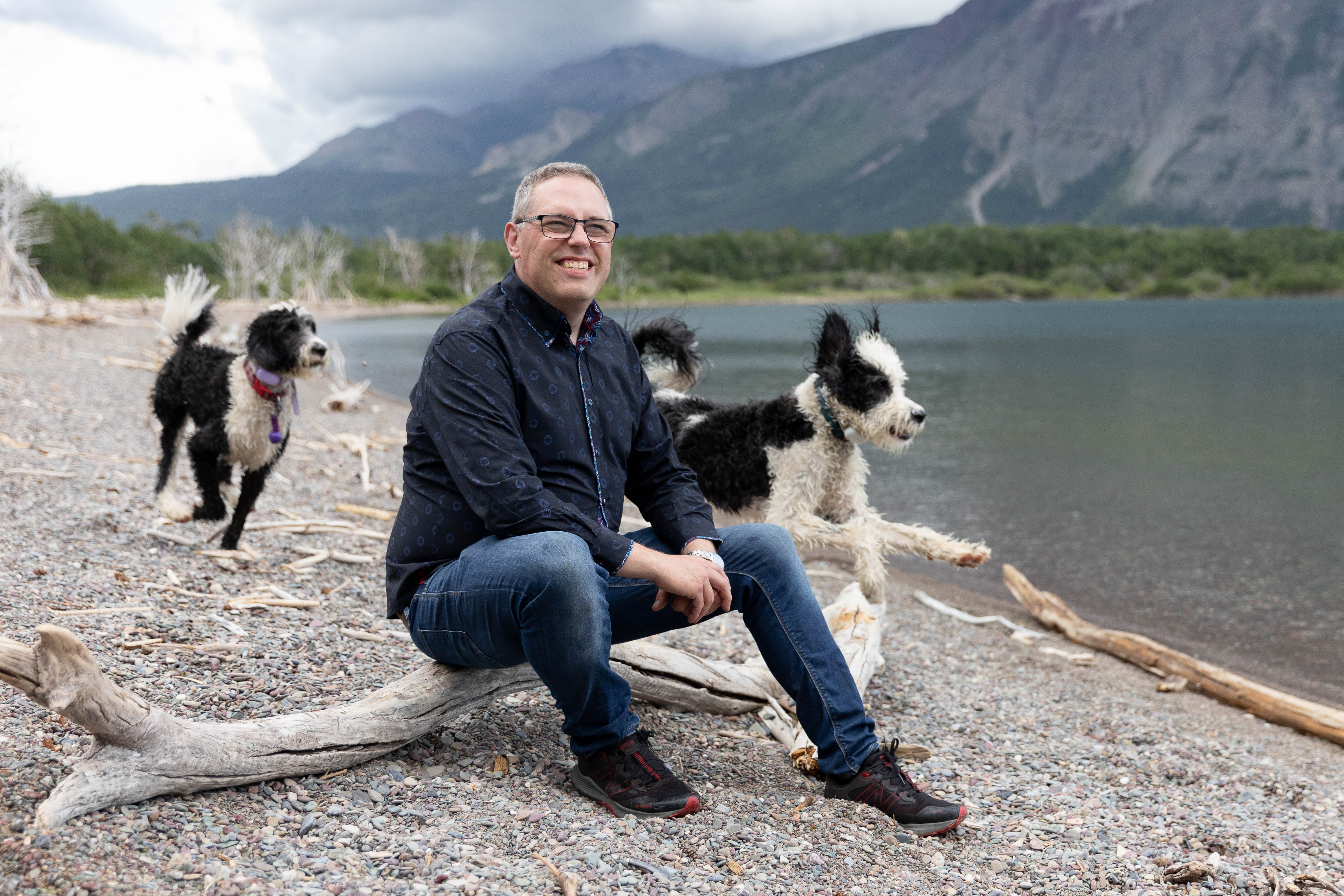 Peter sitting on a beach in Waterton National Park while his Sheepadoodles Walter and Maggie run by him